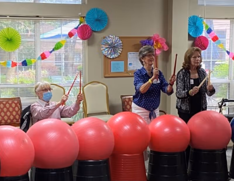 Three elderly women participating in a group activity indoors, playing percussion instruments on large red exercise balls placed on black stands. The room is decorated with colorful paper decorations and garlands, and there are chairs and windows in the background.
