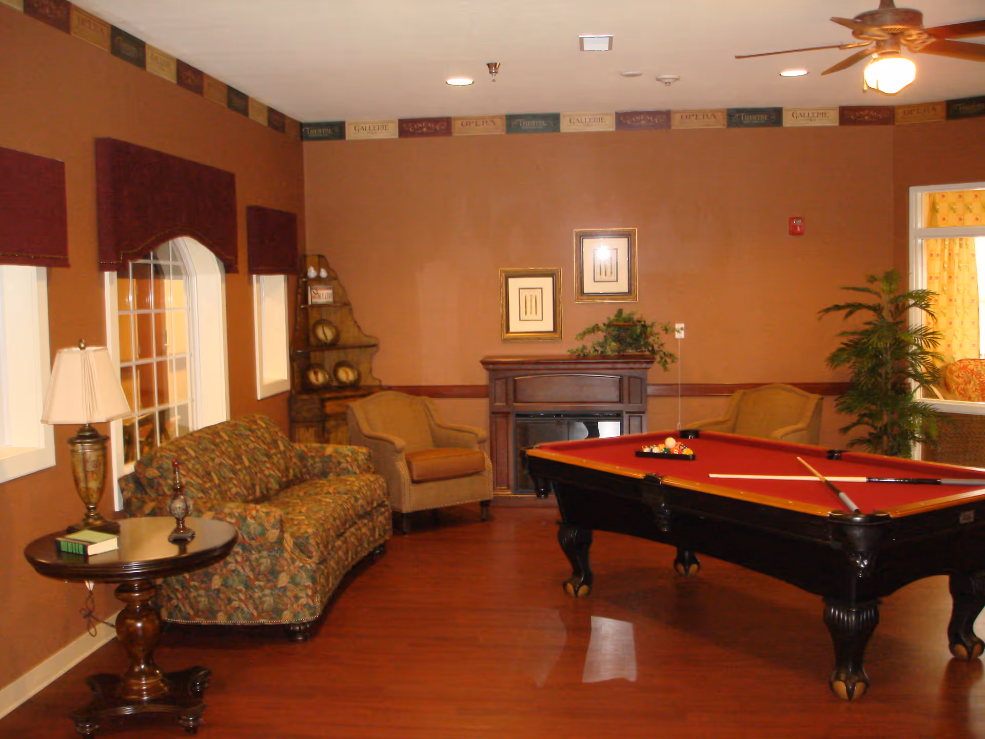 A warm-toned lounge with a red-felt pool table, floral sofa, armchairs, and a decorative fireplace.
