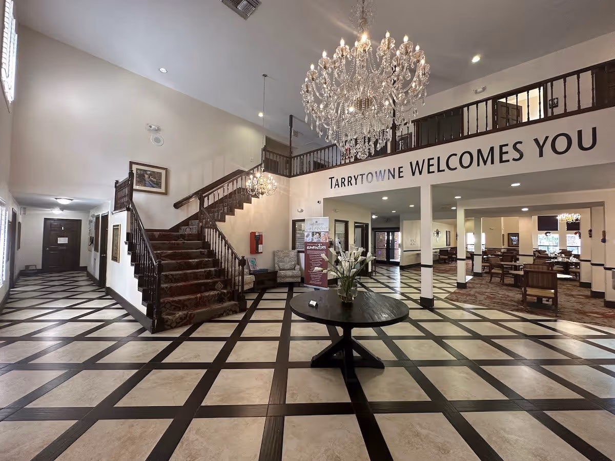 Spacious and elegant lobby area of Tarrytowne Estates featuring a large chandelier, a round table with a flower arrangement, a carpeted staircase with wooden railings, and a welcoming sign that reads 'Tarrytowne Welcomes You'. The floor has a checkered pattern, and there are seating areas and a dining room visible in the background.