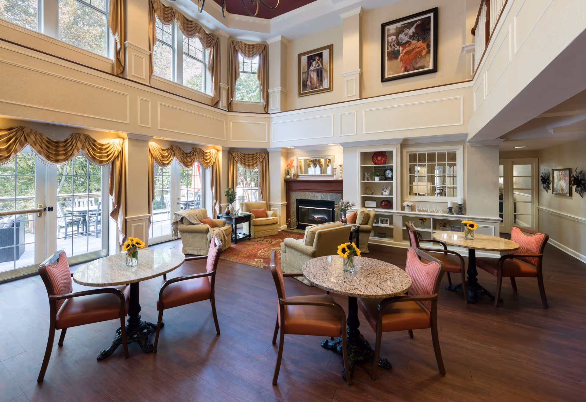 A spacious and well-lit senior living common area with high ceilings and large windows. The room features round tables with chairs, each table adorned with a small vase of sunflowers. Comfortable armchairs and a fireplace with a mantel are arranged near the back wall. The walls are decorated with framed artwork and built-in shelves holding decorative items. Large windows with elegant drapes allow natural light to fill the room, and a door leads to an outdoor patio area with seating.