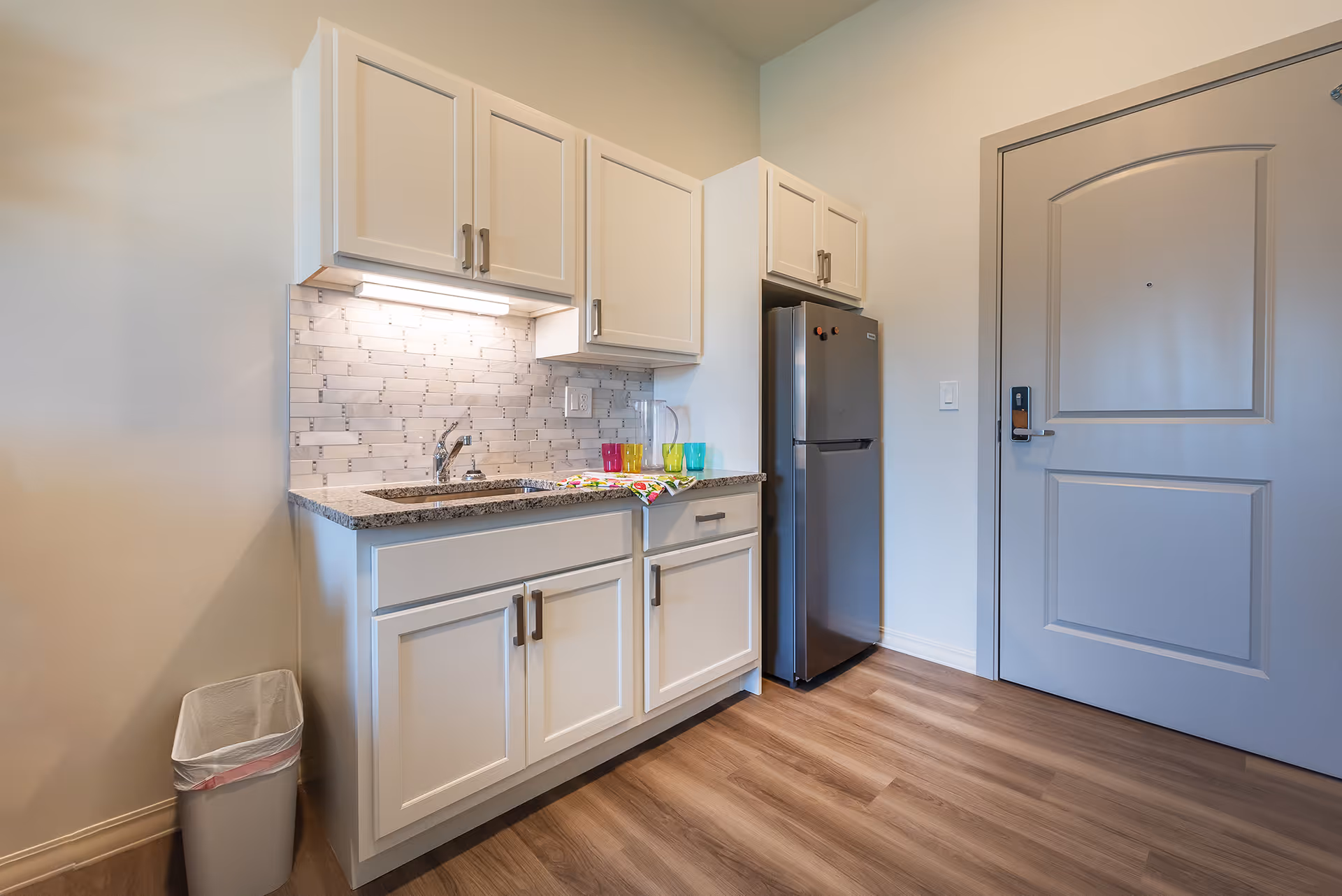 Compact kitchenette with white cabinets, a sink under a tiled backsplash, a small stainless refrigerator and an entry door.