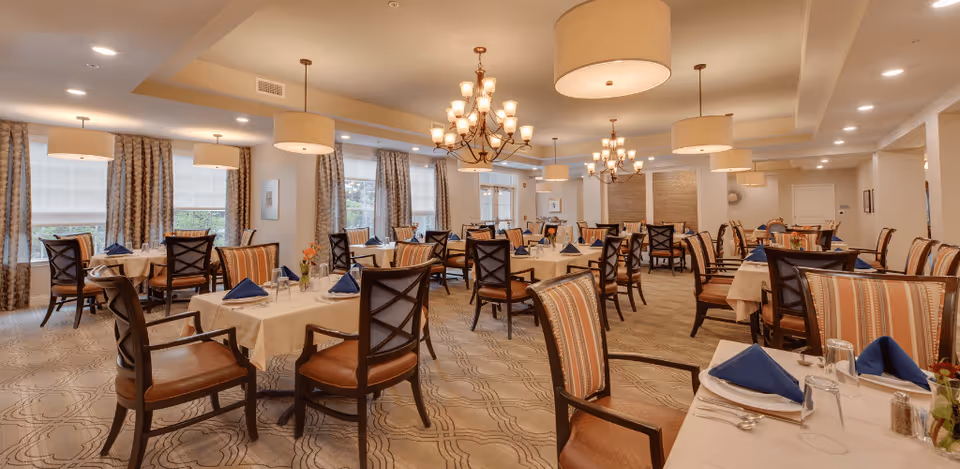 A spacious dining room with multiple tables covered in white tablecloths, each set with blue folded napkins, plates, glasses, and silverware. The room features patterned carpet, large windows with curtains, and warm lighting from chandeliers and ceiling lamps. Chairs with wooden frames and striped cushions surround the tables.