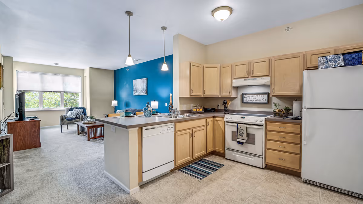Open kitchen and living room area in a senior living facility. The kitchen features light wood cabinets, a white refrigerator, stove, dishwasher, and a countertop with two pendant lights hanging above. The living room has a blue accent wall, a window with blinds, a TV on a wooden stand, a chair, and a coffee table.