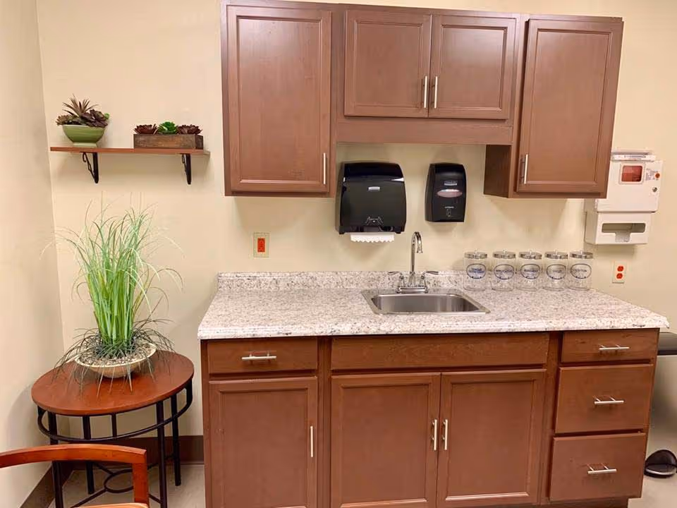 A small kitchenette area with brown wooden cabinets above and below a granite countertop. There is a stainless steel sink with a faucet in the center of the countertop. Mounted on the wall above the sink are a black paper towel dispenser and a soap dispenser. To the right, there are six clear jars with labels on the countertop and a white wall-mounted dispenser. On the left side, there is a small round wooden table with a potted green plant on it, and a wooden chair partially visible. A small wooden shelf with potted succulents is mounted on the wall above the table.