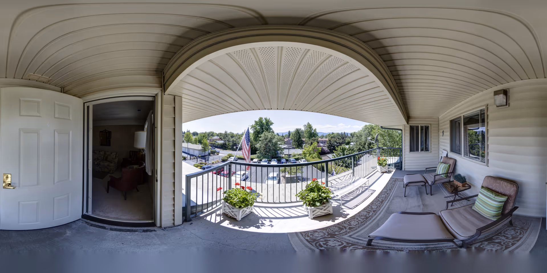 Covered second-floor balcony with lounge chairs, potted flowers, a railing and an open door into the apartment overlooking the neighborhood.