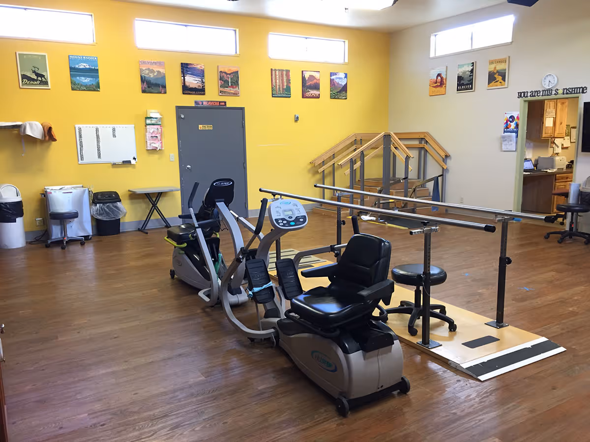 A rehabilitation room with exercise equipment including a recumbent bike and parallel bars for walking practice. The room has yellow walls decorated with landscape posters and a wooden floor. There is a small table, trash bins, and a doorway leading to another room with kitchen cabinets visible.