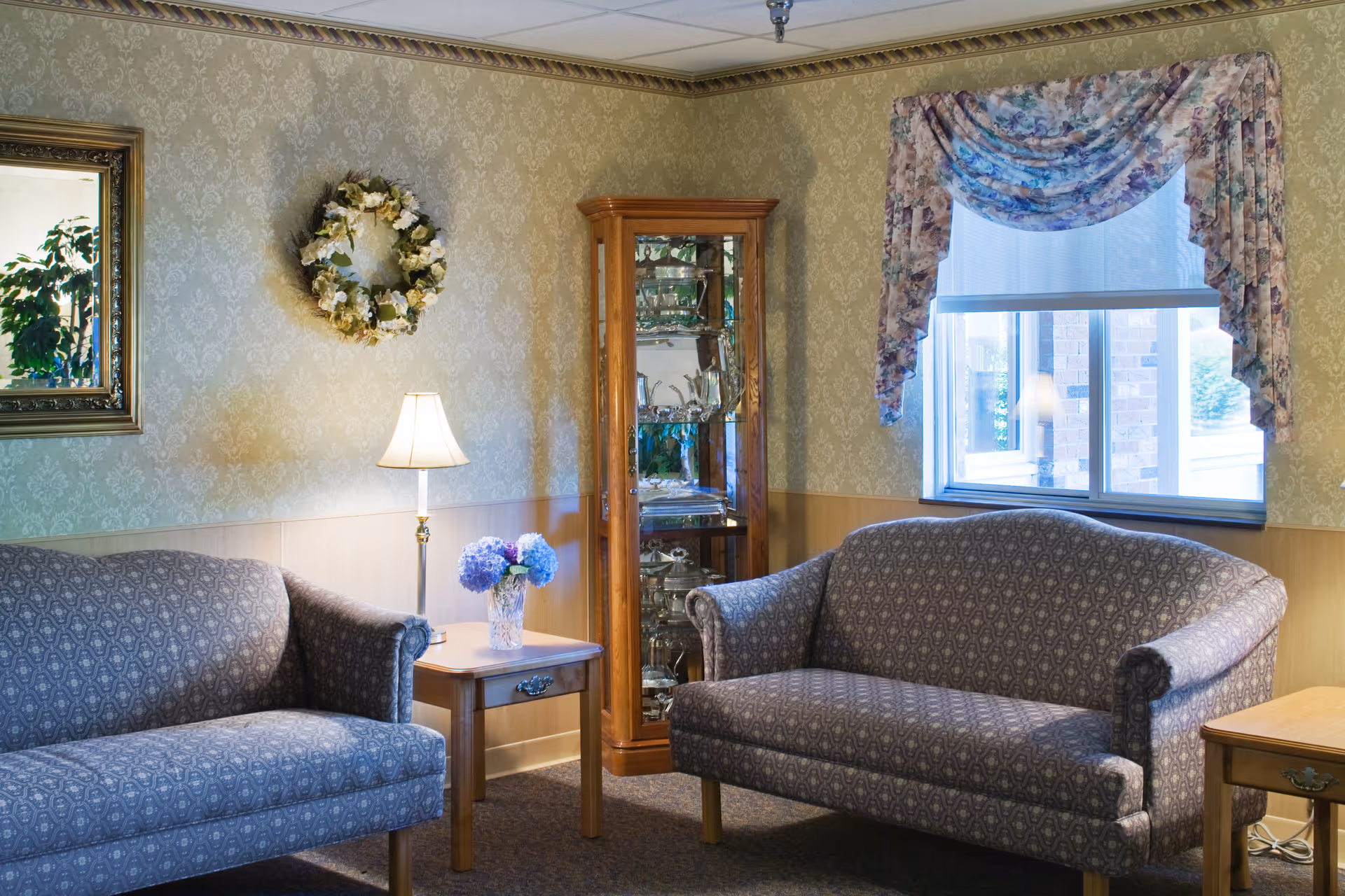 A cozy living room area with two patterned upholstered sofas facing each other, a wooden side table with a lamp and a vase of blue flowers, a wooden display cabinet with glass shelves holding decorative items, a floral wreath on the wallpapered wall, a large framed mirror, and a window with floral curtains letting in natural light.