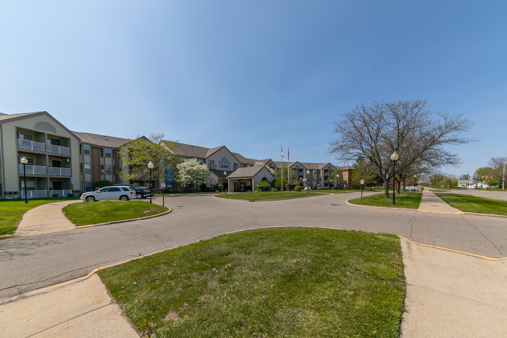 Front exterior of a multi-story senior living building with a circular driveway, flagpoles, parked cars, and landscaped lawns.