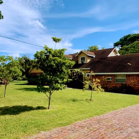 A single-story brick building with a dark shingled roof surrounded by a well-maintained grassy lawn with several small trees under a partly cloudy blue sky.