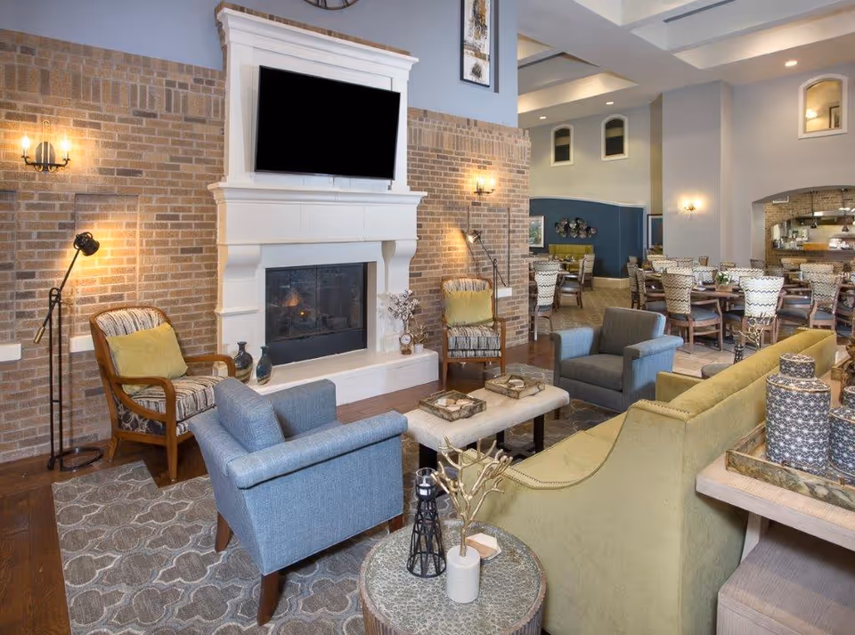 A cozy living room area with a large white fireplace set against a brick wall. Above the fireplace is a mounted flat-screen TV. The room features a mix of seating including a green sofa, two blue armchairs, and two wooden armchairs with patterned cushions and yellow pillows. There are floor lamps and wall sconces providing warm lighting. In the background, there is a dining area with multiple tables and chairs, and a kitchen or serving area visible through an archway.