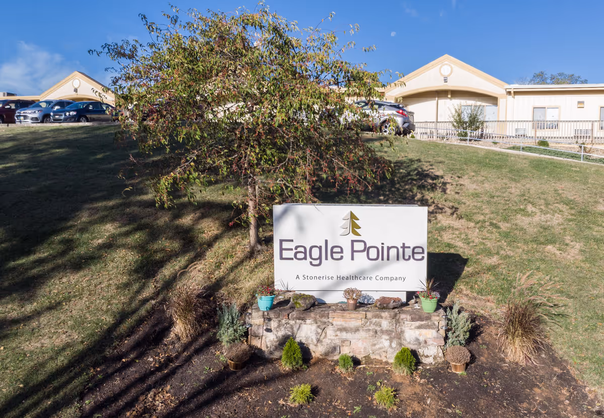 Outdoor view of Eagle Pointe Healthcare Center sign with a tree and small plants around it, parked cars and building structures in the background under a clear blue sky.