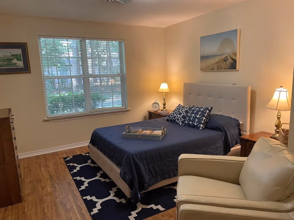 A cozy bedroom with a large window letting in natural light. The room features a bed with a tufted headboard, navy blue bedding, and decorative pillows with anchor and circle patterns. There are two wooden nightstands with lamps on either side of the bed, a beige armchair in the foreground, a framed picture on the wall above the bed, and a wooden dresser to the left. The floor is hardwood with a navy and white patterned rug under the bed.