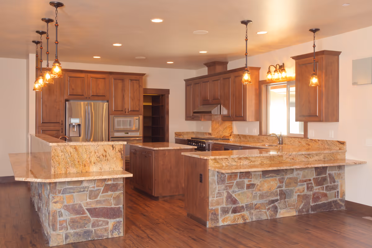 A spacious kitchen with wooden cabinets, granite countertops, and a stone-faced island. The kitchen features stainless steel appliances including a refrigerator and microwave, pendant lighting above the island, and a window above the sink allowing natural light in.