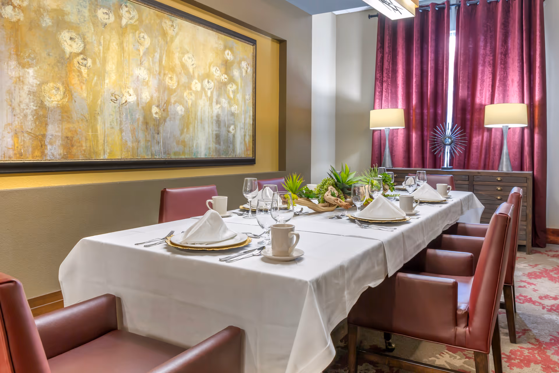 A dining room with a long table covered in a white tablecloth, set with plates, napkins, glasses, cups, and silverware. The table is surrounded by maroon leather chairs. In the background, there is a sideboard with two lamps and decorative items, and a large window with maroon curtains. A large abstract painting with yellow and white tones hangs on the wall.