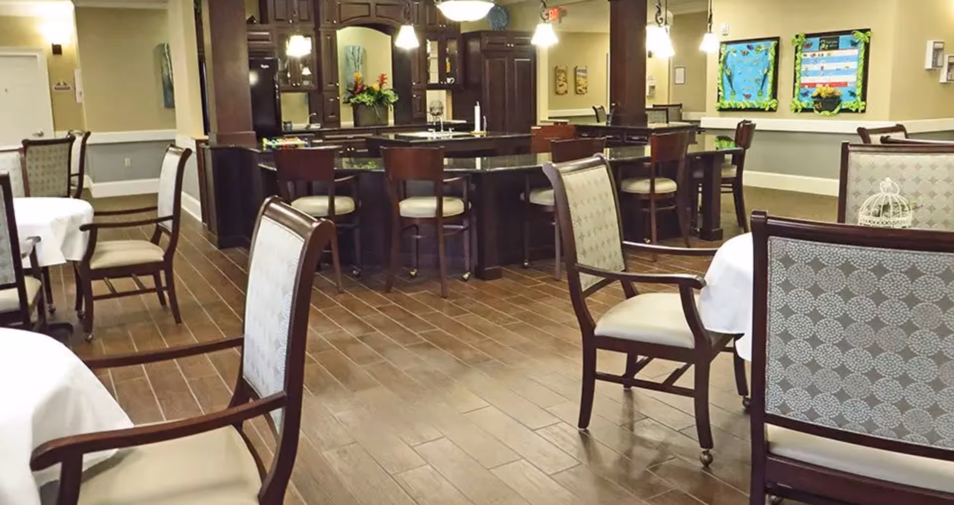 Interior view of a dining area in a senior living facility with wooden chairs and tables covered with white tablecloths. In the background, there is a dark wood bar or counter with high chairs and decorative flowers. The room has wooden flooring and soft lighting, with bulletin boards on the far wall.