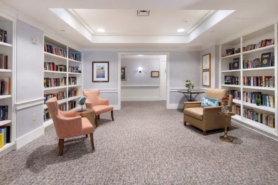 A cozy reading room with built-in white bookshelves filled with books on both sides. The room has a beige carpet and soft lighting from ceiling fixtures. There are two pink upholstered armchairs with a small round table between them on the left side, and a single tan leather armchair with a blue patterned pillow and a small round side table holding a vase with flowers on the right side. The walls are light-colored with framed artwork, and there is an open doorway leading to another room.