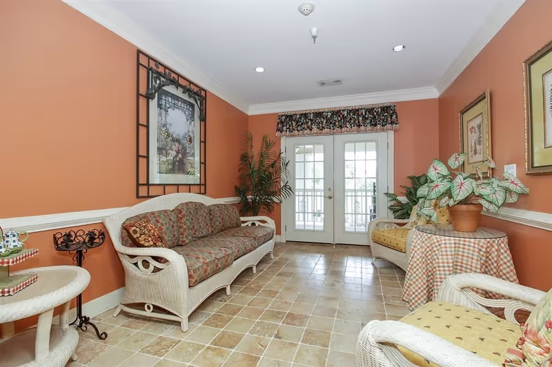 A cozy sitting area with wicker furniture including a sofa and chairs with patterned cushions. The walls are painted a warm terracotta color with white trim. There are framed pictures on the walls, a potted plant on a table with a checkered tablecloth, and a large plant in the corner near double glass doors with a floral valance above them. The floor is tiled with a light stone pattern.