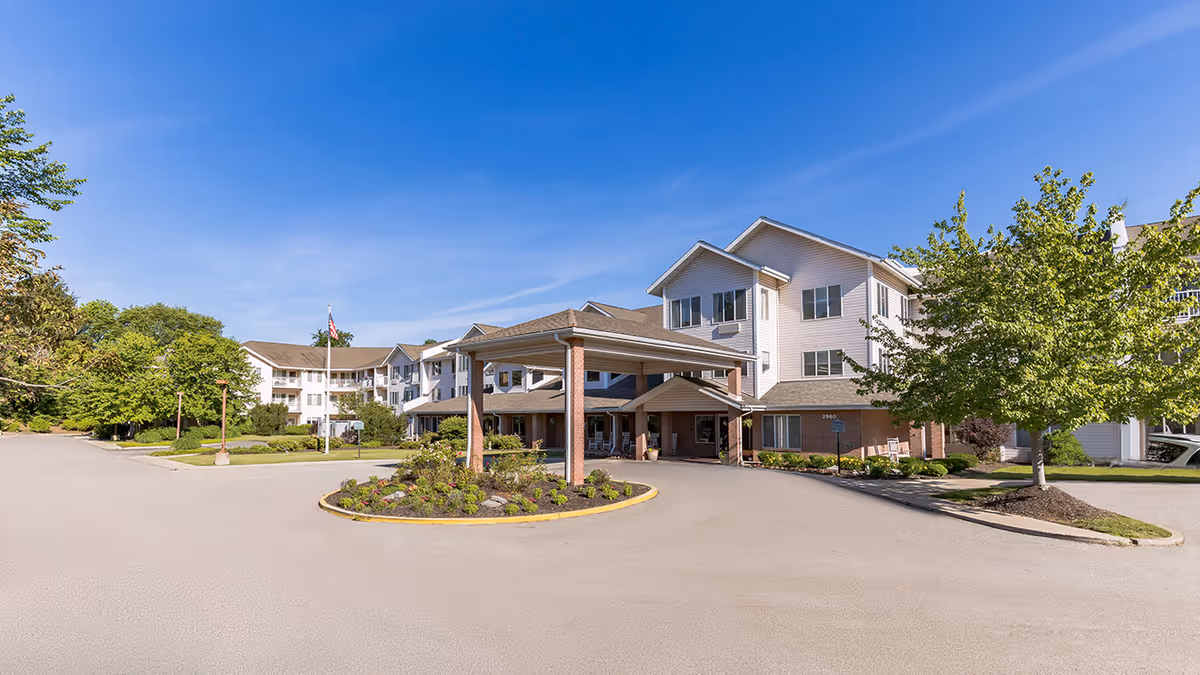 Exterior view of a multi-story senior living facility with a covered entrance, surrounded by trees and landscaping under a clear blue sky.