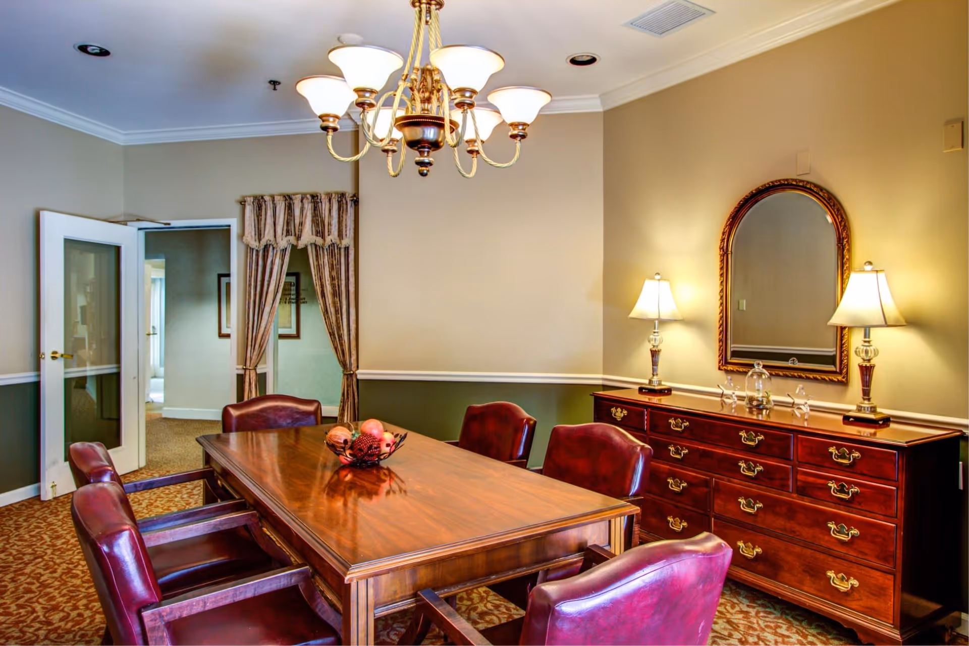 A formal dining room with a polished wooden table surrounded by six burgundy leather chairs. A decorative bowl with fruit sits in the center of the table. The room features a wooden sideboard with brass handles, topped with two lamps and an ornate framed mirror. A chandelier with multiple lights hangs above the table. The walls are painted beige with a white chair rail and green lower panel, and a door with glass panels is open to an adjoining room.