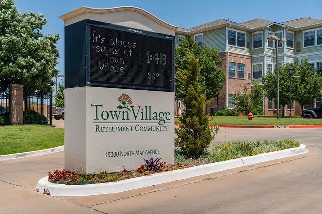 Outdoor view of the entrance sign for Town Village Retirement Community at 13000 North May Avenue, with a digital display showing the message 'It's always sunny at Town Village!' and the temperature 98°F. The background includes a multi-story residential building, trees, and a clear sky.