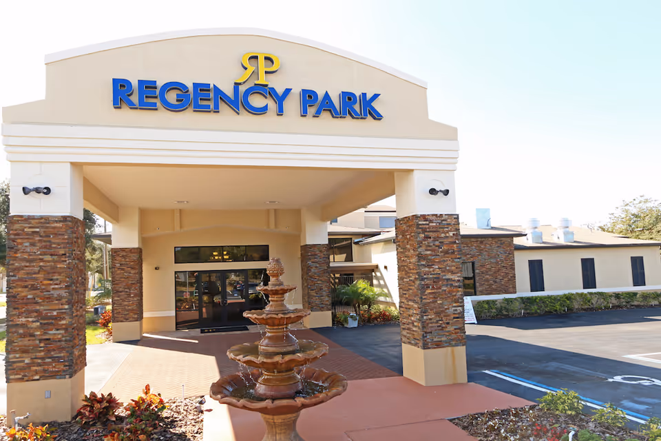 Front exterior view of Regency Park Assisted Living & Memory Care building with a covered entrance supported by stone pillars and a three-tiered water fountain in front.