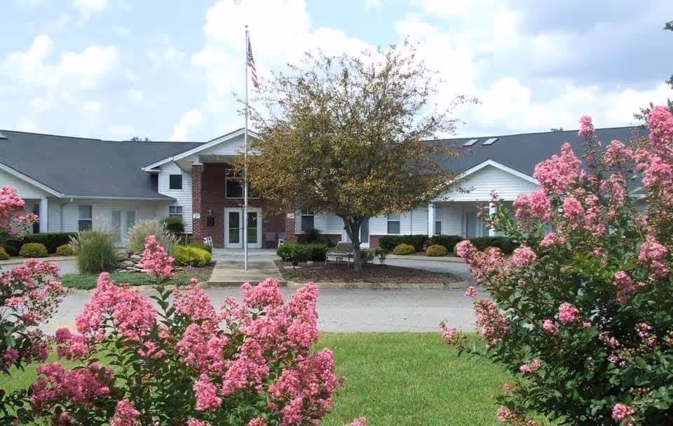 Front exterior view of a single-story assisted living facility building with white siding and a dark roof. There is a flagpole with an American flag in front of the entrance, surrounded by landscaped bushes and a tree. Pink flowering bushes frame the foreground, and the sky is partly cloudy.