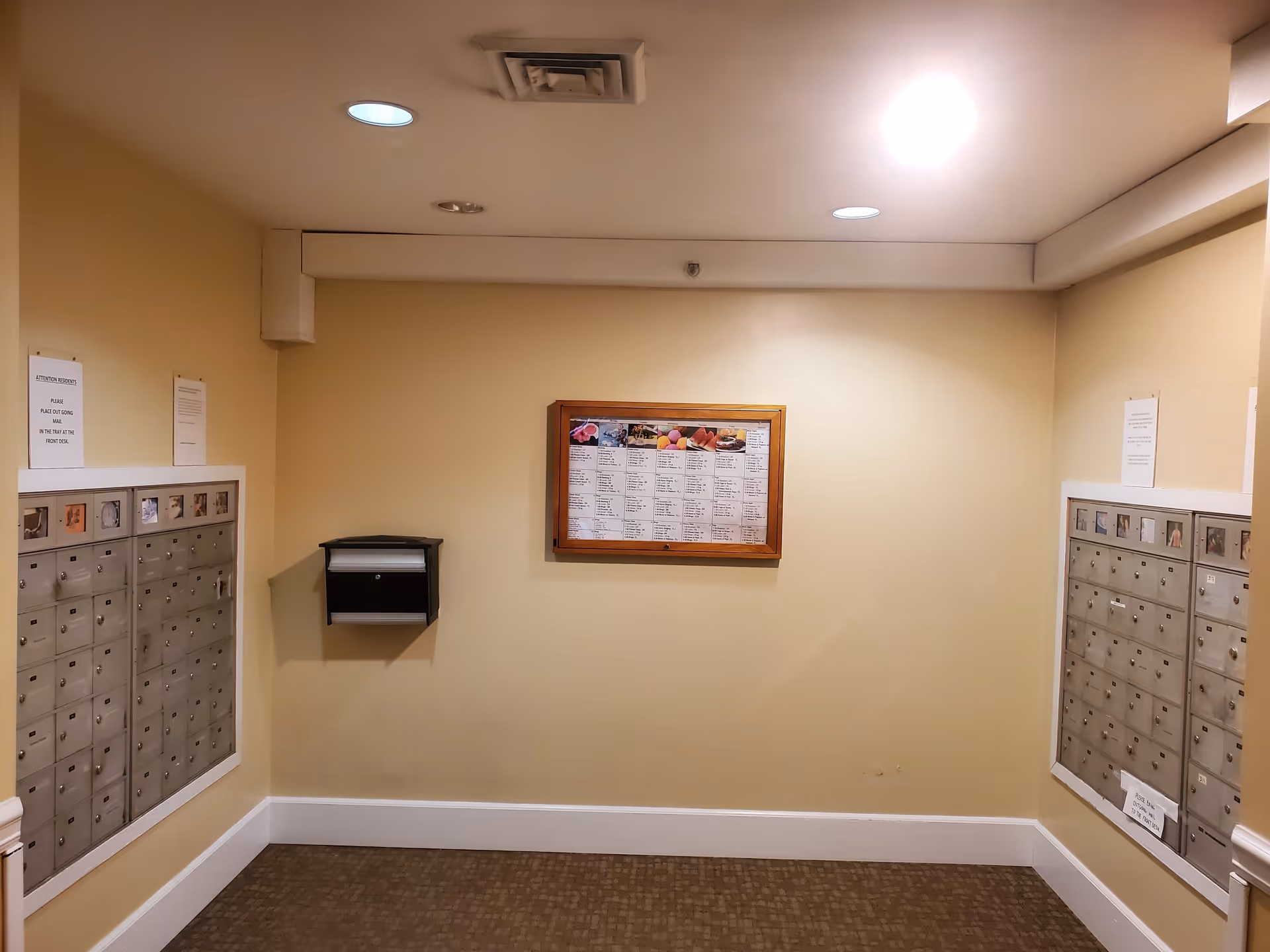 Interior view of a mailroom with two sets of metal mailboxes mounted on opposite walls. A bulletin board with a posted menu is centered on the back wall. The walls are painted beige, and the floor is carpeted. Ceiling lights illuminate the space.