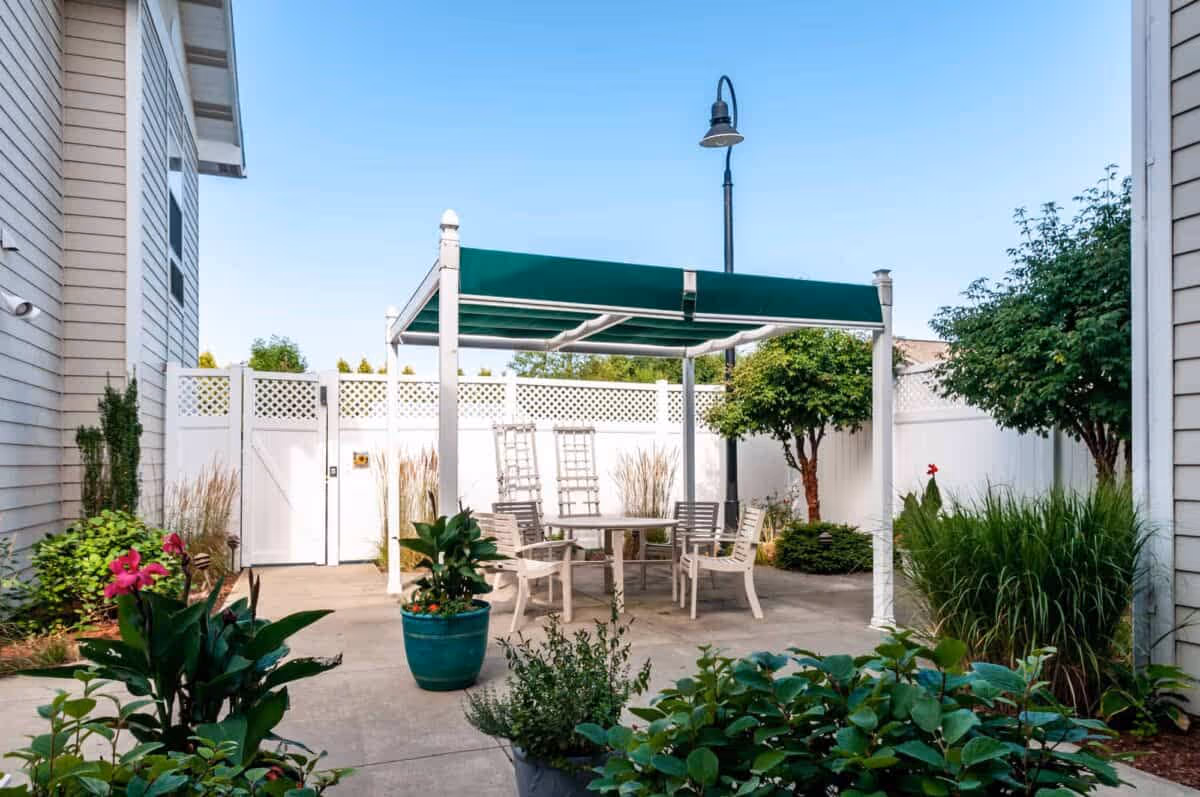 Outdoor patio area with a green canopy over a round table surrounded by white chairs, potted plants, and greenery along a white fence under a clear blue sky.