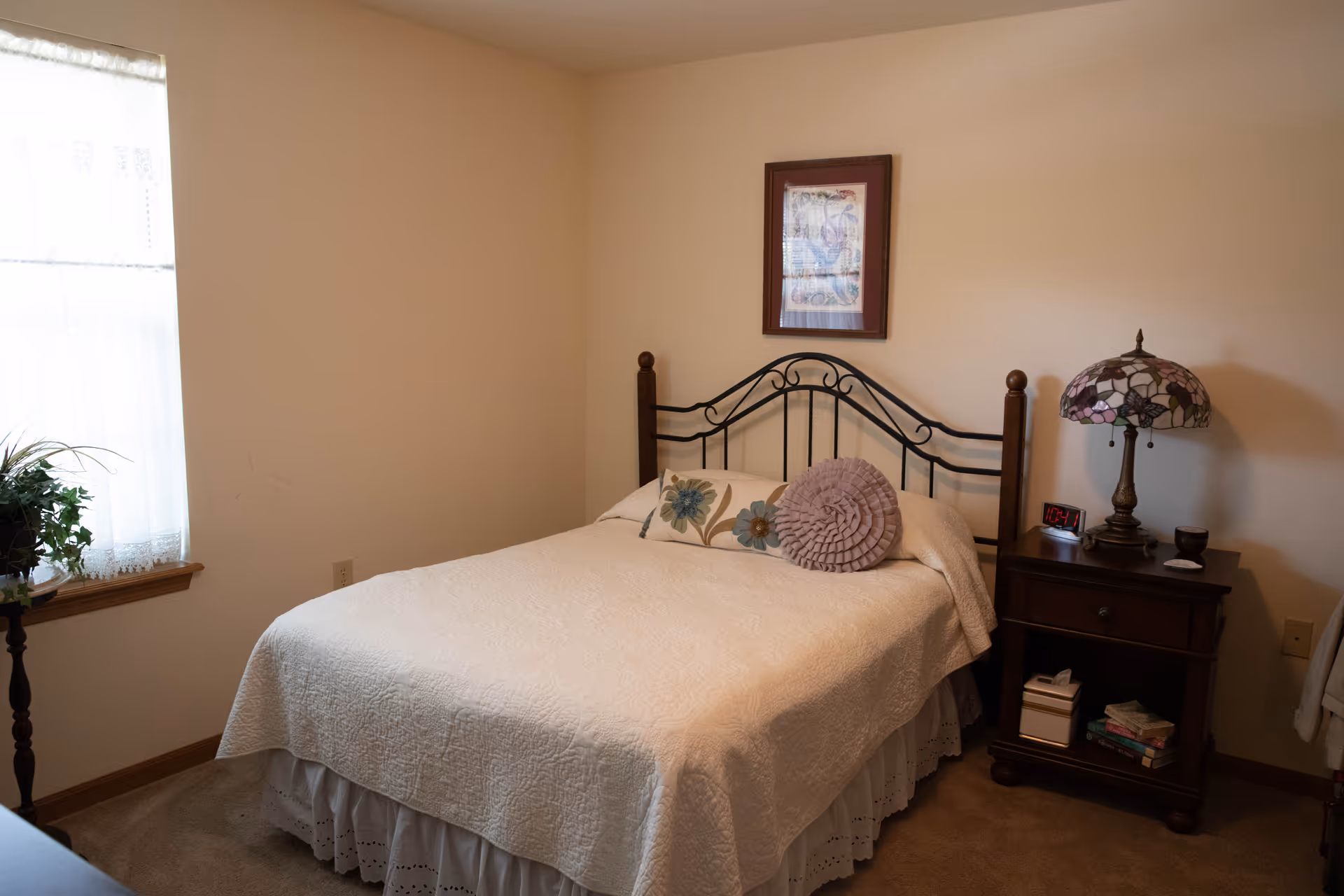A cozy bedroom with a bed covered in a white quilt and decorative pillows. There is a wooden nightstand with a Tiffany-style lamp, a digital clock, and some small items. A framed picture hangs on the beige wall above the bed. A window with sheer white curtains and a plant on a stand are visible to the left.