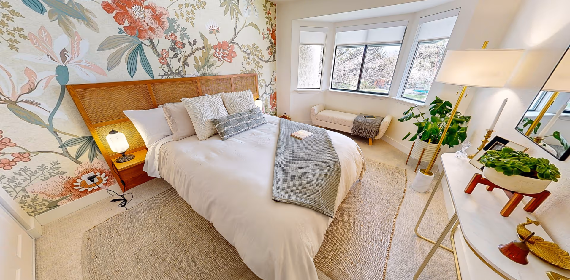 A cozy bedroom with a large bed featuring white bedding and decorative pillows. The wall behind the bed has a floral wallpaper design. There is a small bedside table with a lamp on the left side. A beige bench is placed under a set of three large windows letting in natural light. On the right side, there is a white console table with a potted plant, a lamp, and decorative items. The room has a light-colored carpet and a woven rug under the bed.