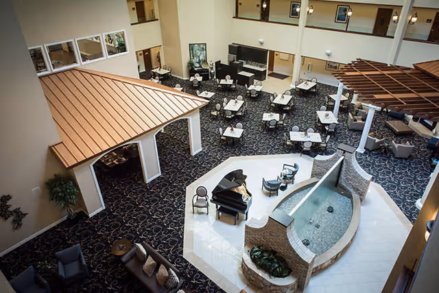 Spacious indoor common area with multiple tables and chairs arranged for dining or socializing, a grand piano with chairs around it, a decorative water feature with a curved glass panel, and comfortable seating areas under a wooden pergola and near a small roofed structure.