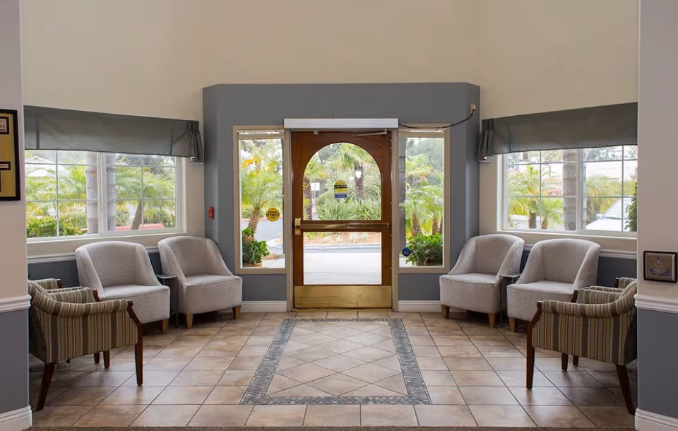 Bright entrance lobby with upholstered chairs arranged on either side of a central glass door and tiled floor, palms visible outside.
