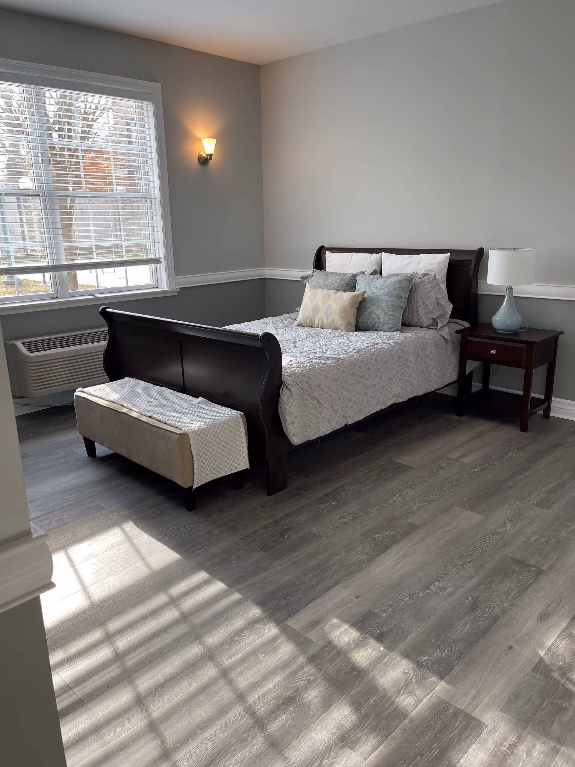 A bright bedroom with a large window letting in natural light. The room features a dark wooden sleigh bed with white and patterned pillows and a light gray bedspread. At the foot of the bed is a beige bench with a white textured throw. A wooden nightstand with a light blue lamp is beside the bed. The walls are painted light gray with white trim, and the floor is covered with gray wood-like planks.
