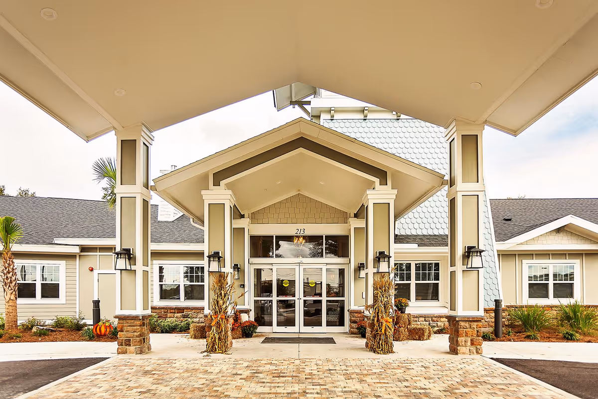 Front entrance of The Canopy at Harper Lake facility featuring a covered driveway with four large pillars decorated with autumn-themed cornstalks and pumpkins. The building has beige siding, stone accents at the base of the pillars, and large glass double doors under a peaked roof.