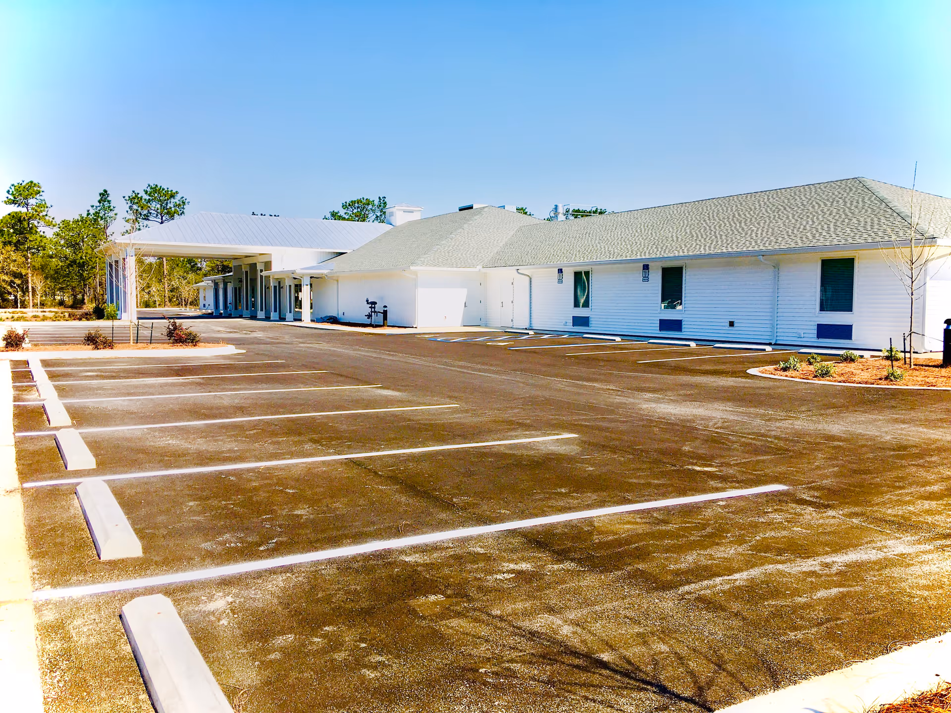 Empty parking lot in front of a single-story white building with a covered entrance and surrounding landscaping.