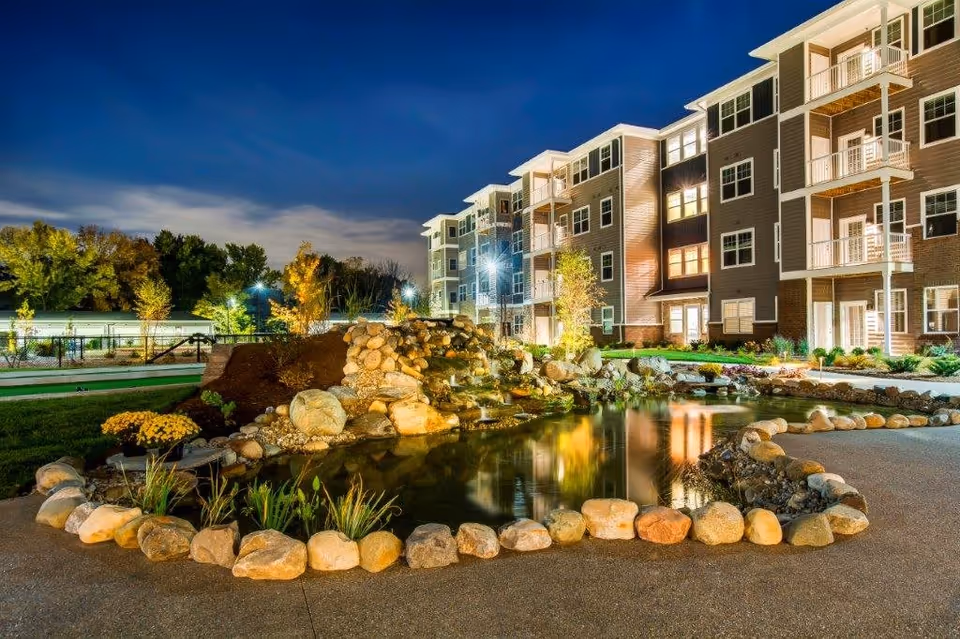 Night view of Encore 55+ Boutique Apartments in Plainfield featuring a well-lit multi-story residential building with balconies. In the foreground, there is a landscaped pond surrounded by rocks and plants, with a small waterfall feature. Trees and additional lighting are visible in the background under a dark blue sky.