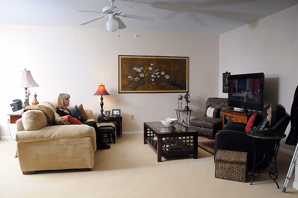 A cozy living room in an assisted living facility with a beige sofa on the left where a woman is seated, a glass coffee table in the center, a patterned armchair and a black recliner on the right, a TV on a wooden stand, two side tables with lamps, and a large floral artwork hanging on the wall.