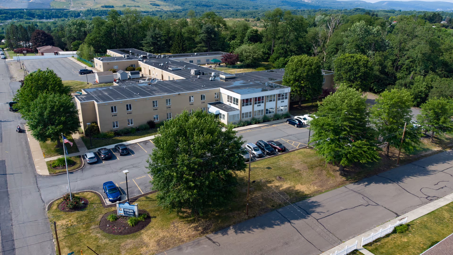 Aerial view of Oak Ridge Rehabilitation and Healthcare Center, a large beige multi-wing building surrounded by trees and parking lots with several cars parked. The facility is located in a green, semi-rural area with hills in the background. There is an American flag and a sign near the entrance.