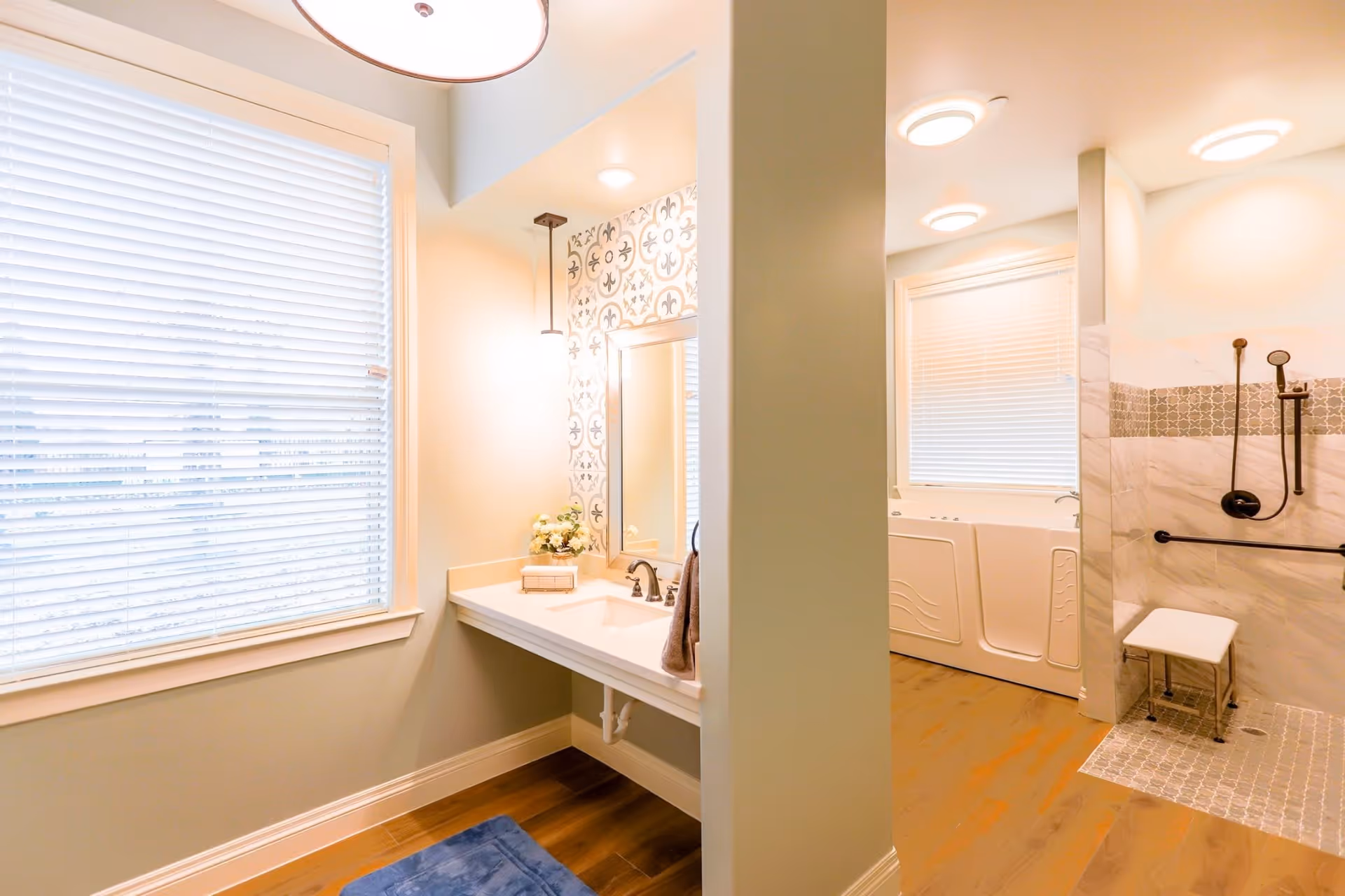 Bright and spacious bathroom with a large window covered by white blinds, a vanity with a sink and mirror, decorative wall tiles, a walk-in shower with a bench and grab bars, and a walk-in bathtub. The floor is wooden with a blue rug near the sink area.