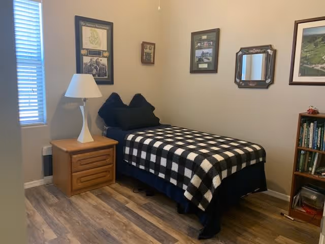 A small bedroom with a single bed covered with a black and white checkered blanket and two black pillows. Next to the bed is a wooden nightstand with a white lamp. The room has light-colored walls decorated with framed pictures and a mirror. There is a window with blinds on the left side and a wooden bookshelf on the right side filled with books and other items. The floor is wood-patterned.