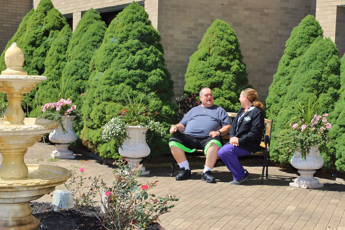 Two people sitting on a bench in a landscaped outdoor courtyard with a fountain, planters, and trimmed evergreen shrubs.