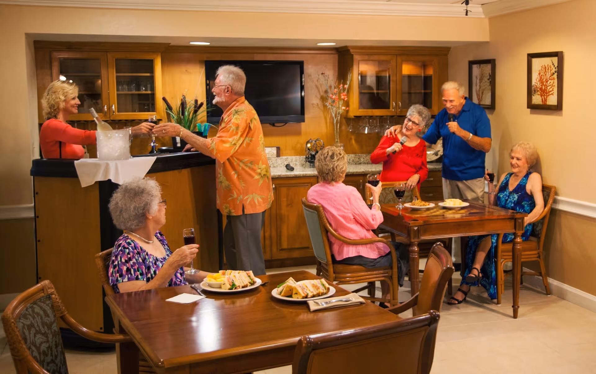A group of elderly people socializing in a cozy room with wooden cabinets and a small bar. Two people are standing at the bar exchanging drinks, while four others are seated at two tables enjoying food and drinks. Two individuals are singing karaoke with microphones. The room has warm lighting and framed artwork on the walls.