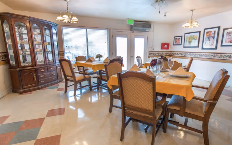 A dining room with several tables covered in yellow tablecloths, each set with plates, glasses, and napkins. There are wooden chairs with cushioned seats around the tables. A large wooden china cabinet filled with glassware is against one wall. The room has framed artwork on the walls, a patterned border trim, and large windows with blinds letting in natural light.