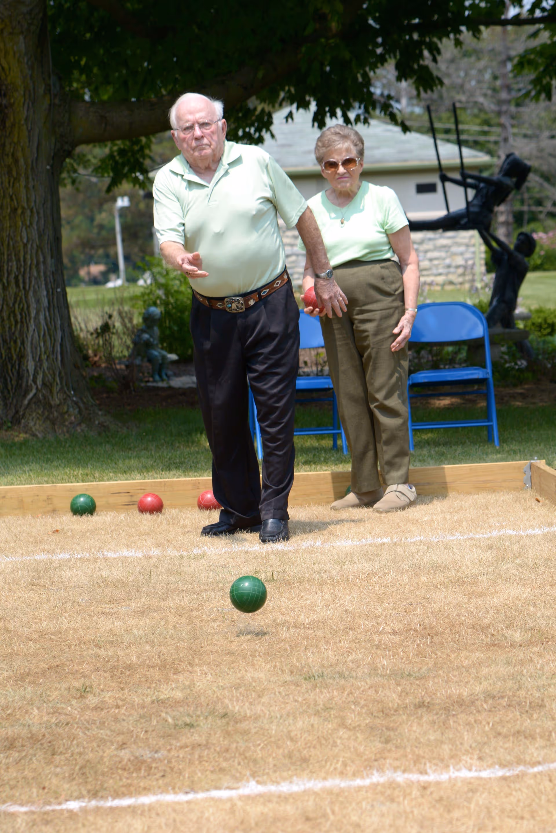 An elderly man and woman playing bocce ball outdoors on a grassy court. The man is in the process of throwing a green bocce ball while the woman watches. There are blue chairs and a tree in the background, along with a sculpture and a building.