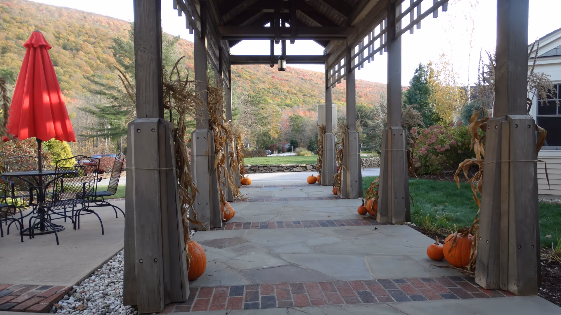 Covered outdoor walkway decorated with pumpkins and dried corn stalks on wooden posts, with a red patio umbrella and metal chairs and tables to the left, and a scenic view of a tree-covered hill in the background.