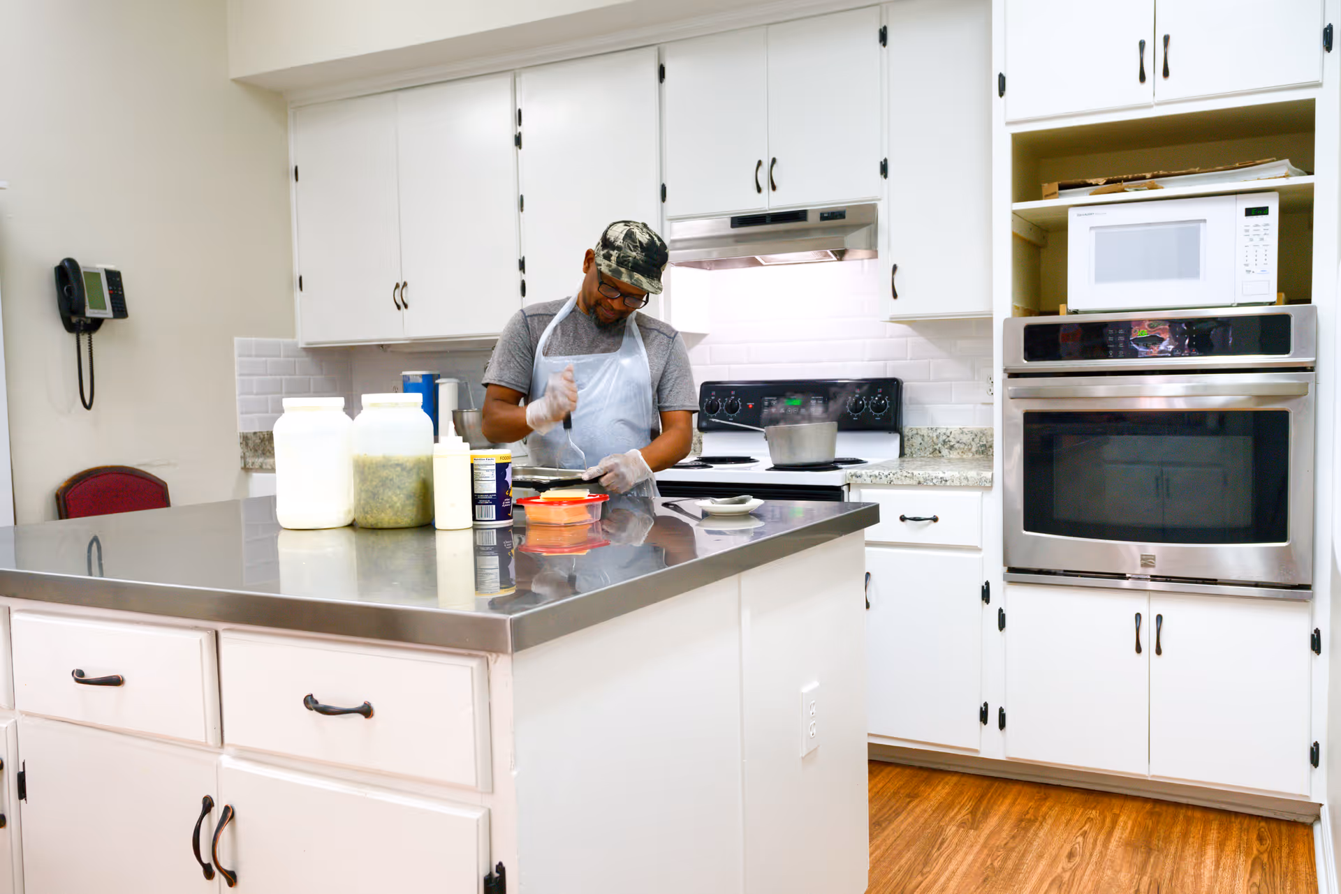 A man wearing a camouflage cap, glasses, gray t-shirt, and apron is preparing food in a kitchen with white cabinets and stainless steel appliances. There are containers and a pot on the kitchen island and stove.