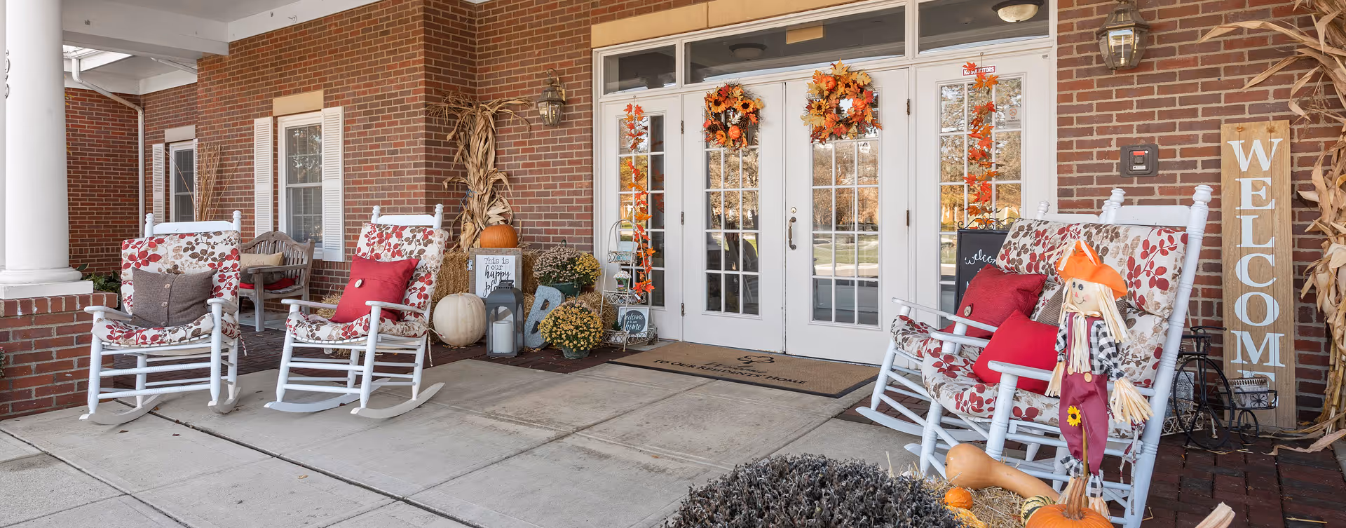 Front entrance of a brick building decorated for autumn with white rocking chairs featuring floral cushions and red pillows, pumpkins, fall wreaths on the glass doors, a scarecrow, and a wooden welcome sign.