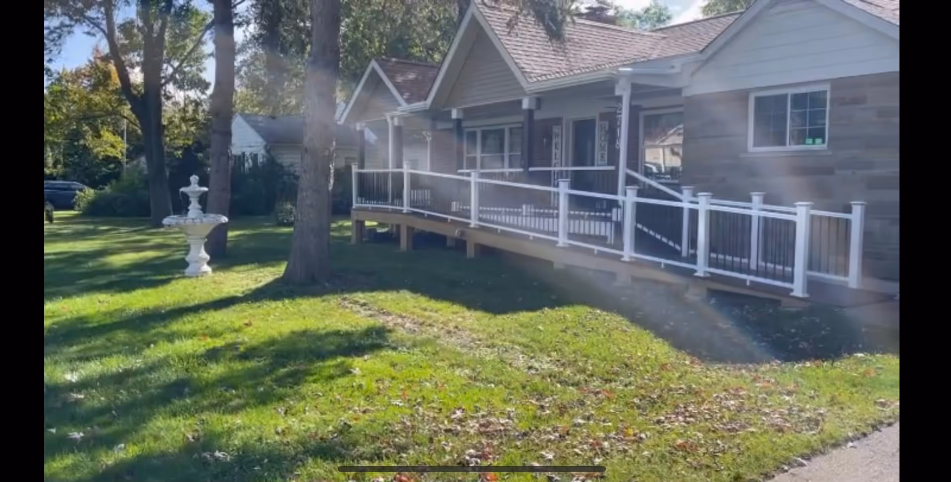 Exterior view of a single-story residential building with a sloped roof and a wheelchair accessible ramp with white railings. The building is surrounded by a green lawn with trees and a white decorative fountain. The scene is sunny with shadows cast by the trees.