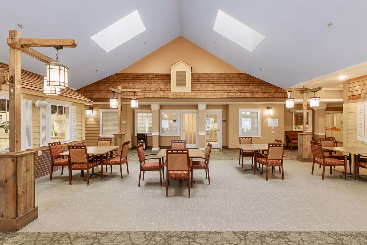 Interior view of a senior living facility common area with multiple wooden tables and chairs arranged neatly. The room features a high vaulted ceiling with skylights, warm lighting fixtures hanging from wooden beams, and walls designed to resemble the exterior of houses with windows and shutters.