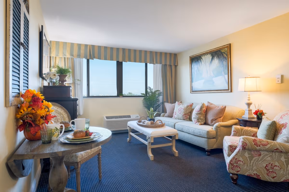 A cozy living room in a senior living facility with a beige sofa adorned with floral and patterned cushions, a matching floral armchair, a white cushioned coffee table, and a small round dining table set with plates, glasses, and a bagel. The room has a large window with striped valance curtains, a framed painting on the wall, a table lamp, and decorative plants and flowers.